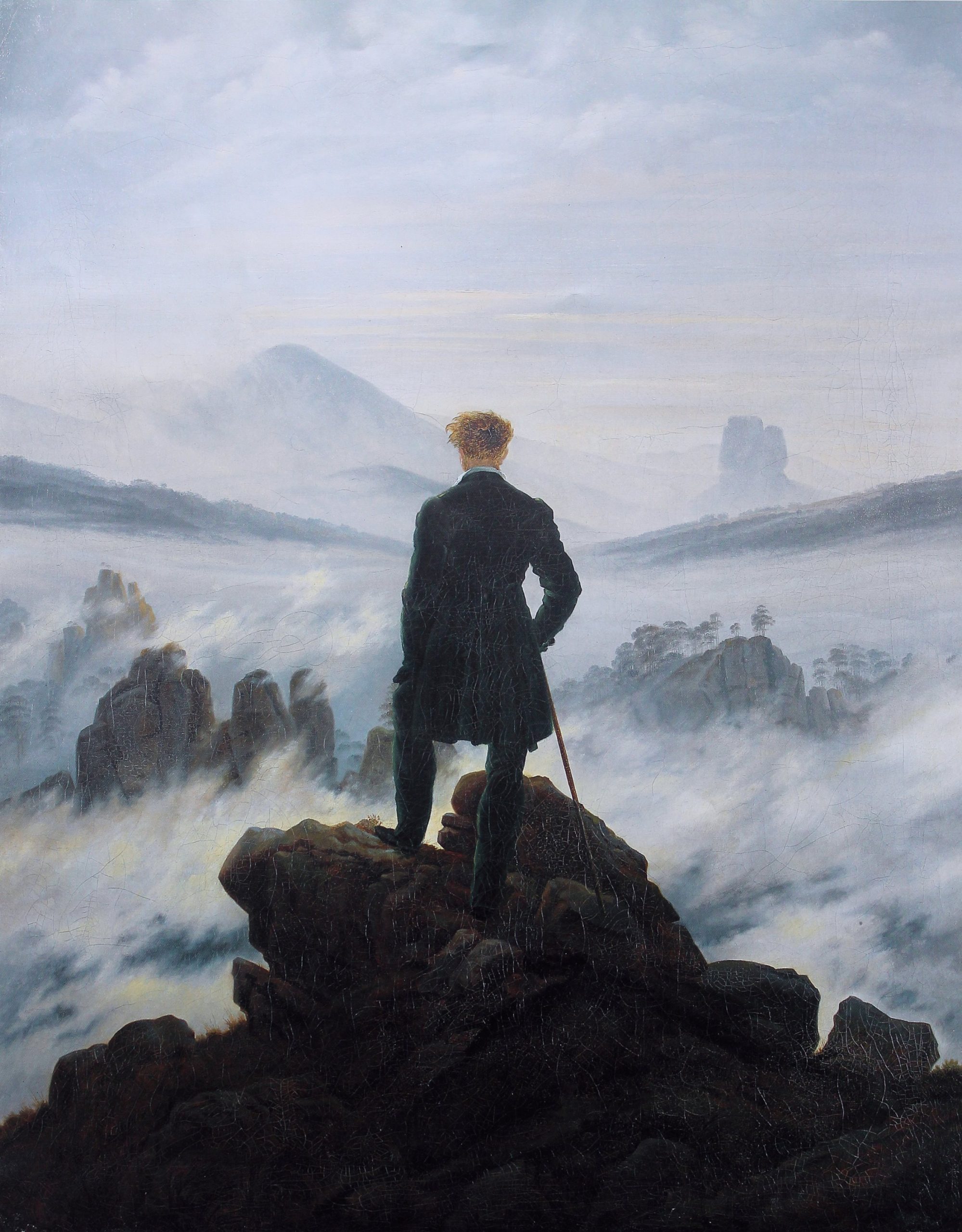 A man whose back is turned towards the viewer standing on a rock before a violent sea
